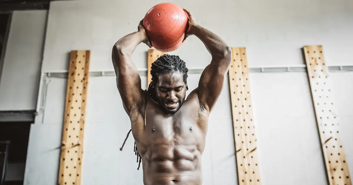 A shirtless man with dreadlocks lifts a red medicine ball overhead in a gym