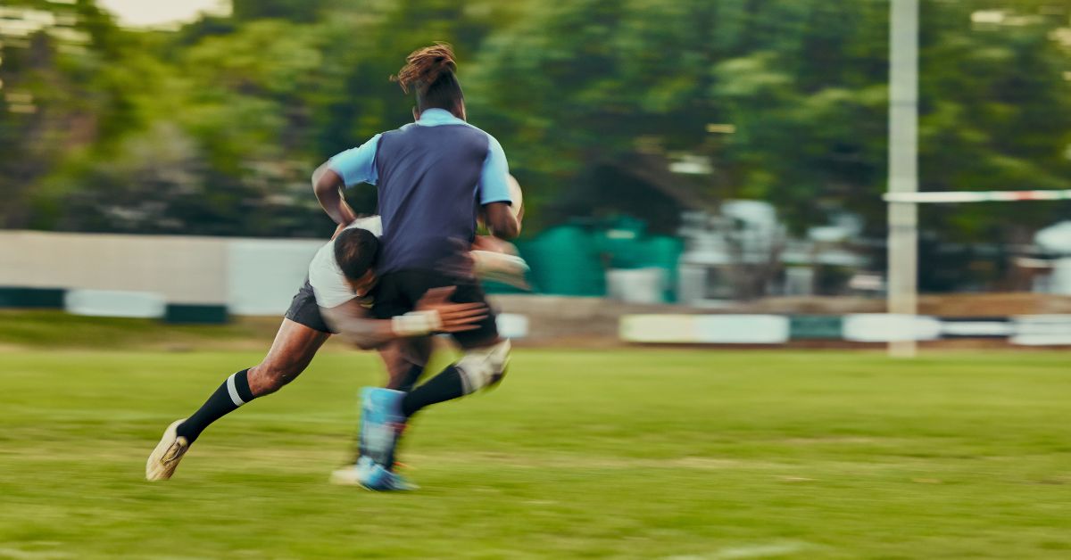 Two rugby players on a field in action. One player is tackling the other. Both are wearing sports gear